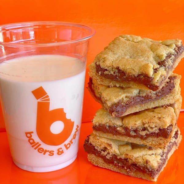 A Ballers & Brew cup of milk next to a stack of chocolate-filled cookie bars on an orange background.