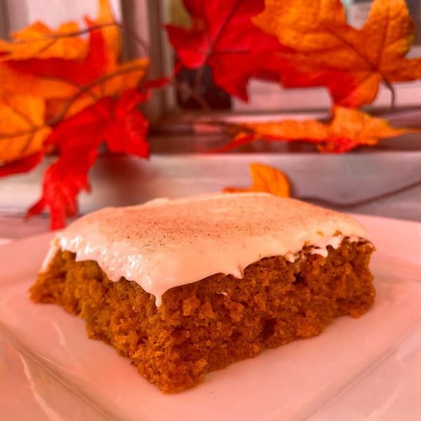 A slice of pumpkin spice cake with white frosting and cinnamon, surrounded by autumn leaves.