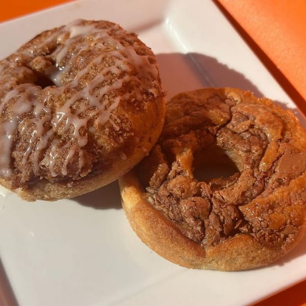Two cinnamon sugar donuts, one with white icing drizzle, on a white plate with an orange background.